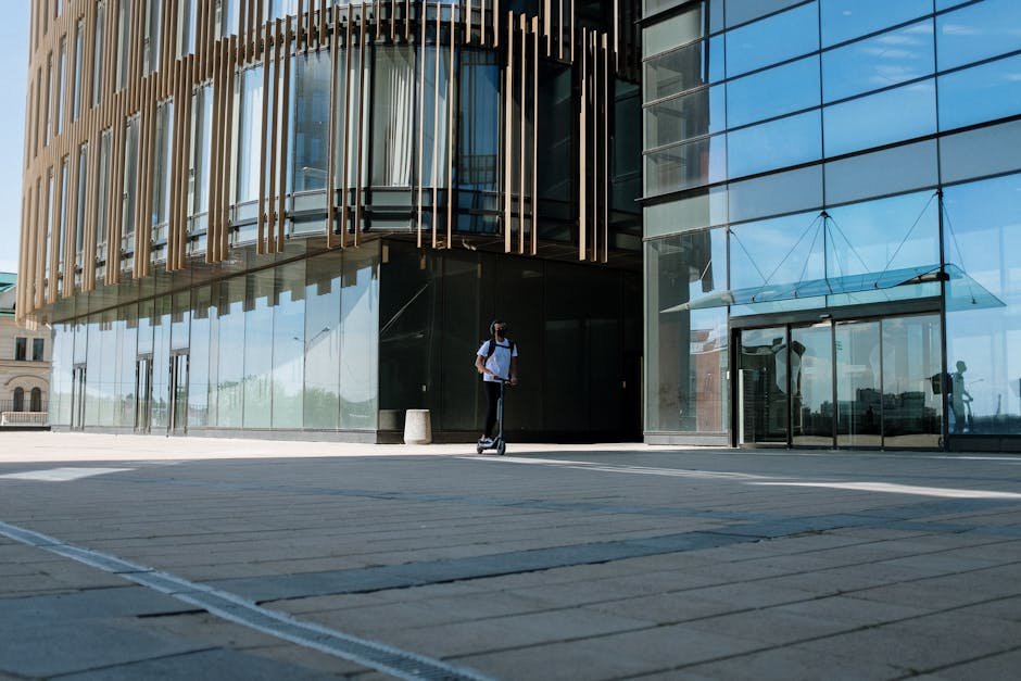 Person riding electric scooter next to a modern office building in the city. Captures urban mobility.