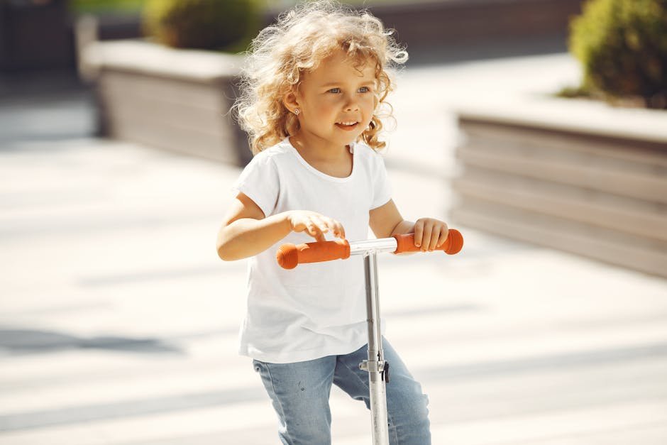 Curly-haired child riding a scooter outdoors on a sunny day, enjoying playtime.
