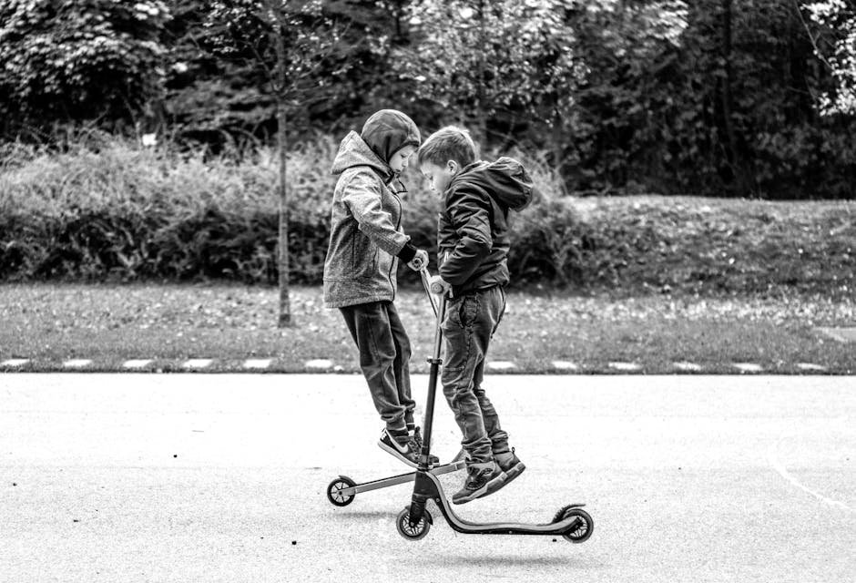 Two young boys playfully jumping on scooters in a monochrome outdoor setting.